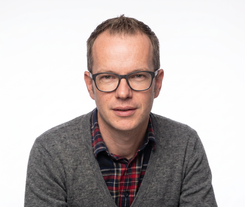 Portrait of a man with short brown hair and dark glasses, wearing a gray cardigan over a red plaid shirt, looking at the camera.